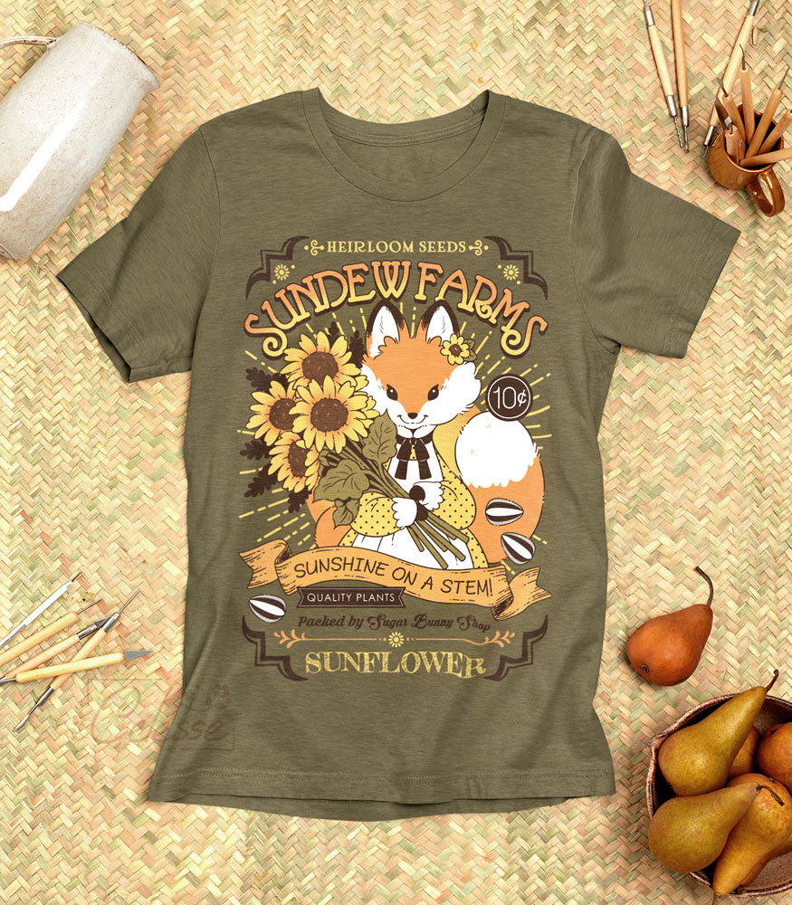 A photo of an olive green shirt laid out on a straw mat, the print featuring a vintage-style logo for Sundew Farms Sunflower Seeds, with a red fox wearing a yellow polka dot dress holding a bundle of sunflowers in their arms.