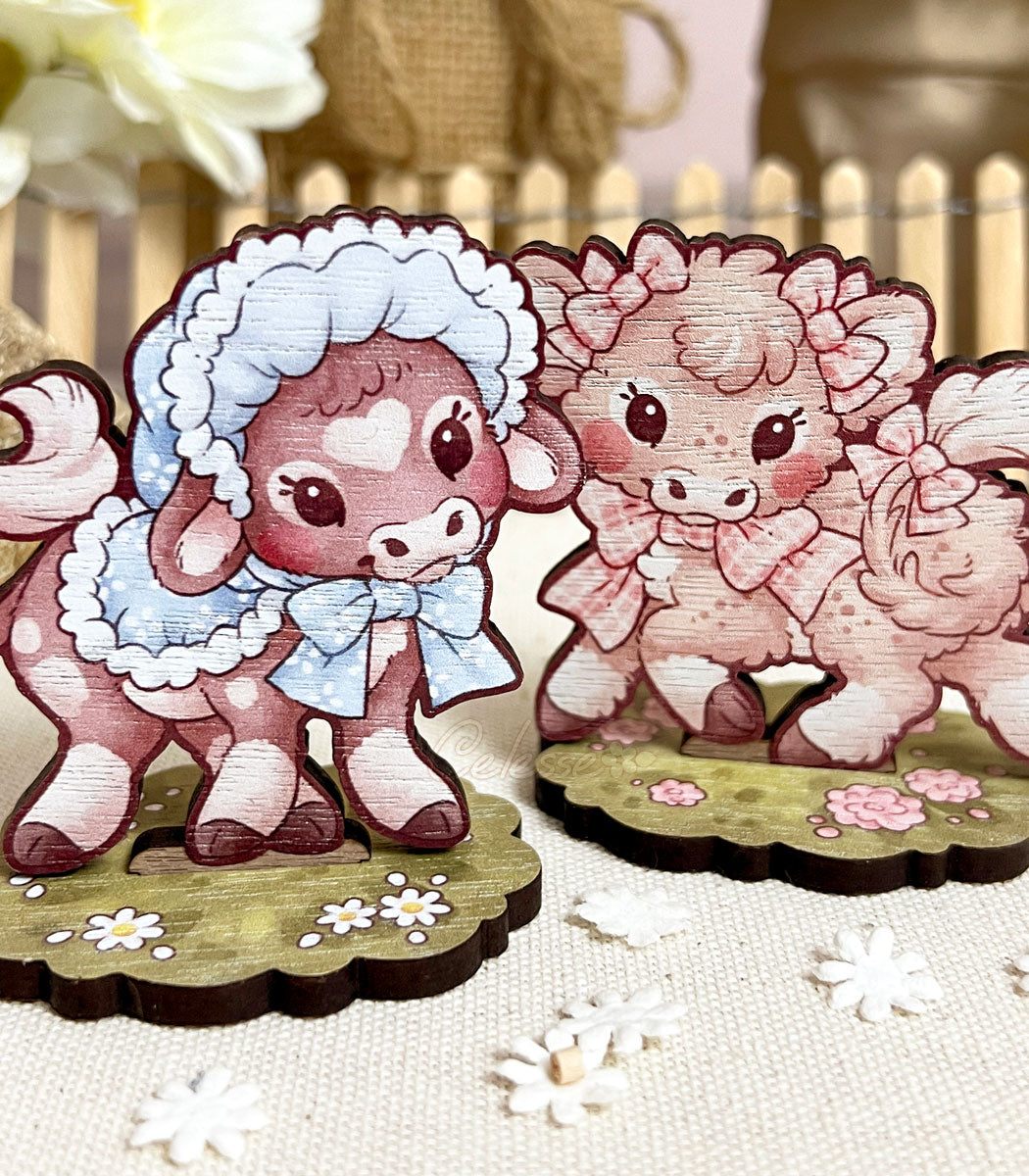 A photo of two wooden standees featuring art of baby cows. The one on the left is dark brown with a heart-shaped spot on their forehead and wears a light blue bonnet and cape. The Right one is a light pinkish-brown color with brown freckles and wears pink gingham ribbons on their ears and tail. Flowers and a picket fence frame them in the background.