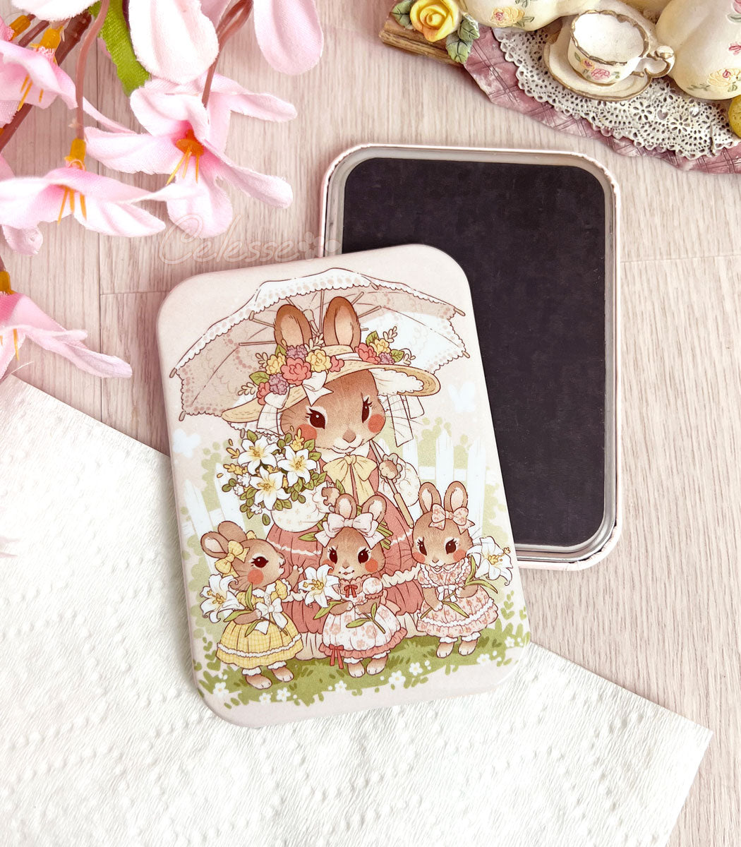 A photo of a rectangular refrigerator magnet featuring artwork of a mother bunny in a vintage style dress holding a bundle of lilies and a lace parasol. Her three daughters stand in front of her each holding their own lily. The back side of a magnet is in the background.