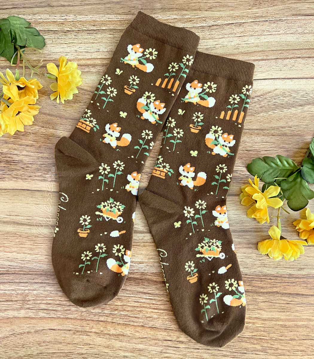 A pair of brown crew socks on a wood surface with flowers, the knit design featuring tiny pixel foxes in vintage clothes frolicking among sunflowers.