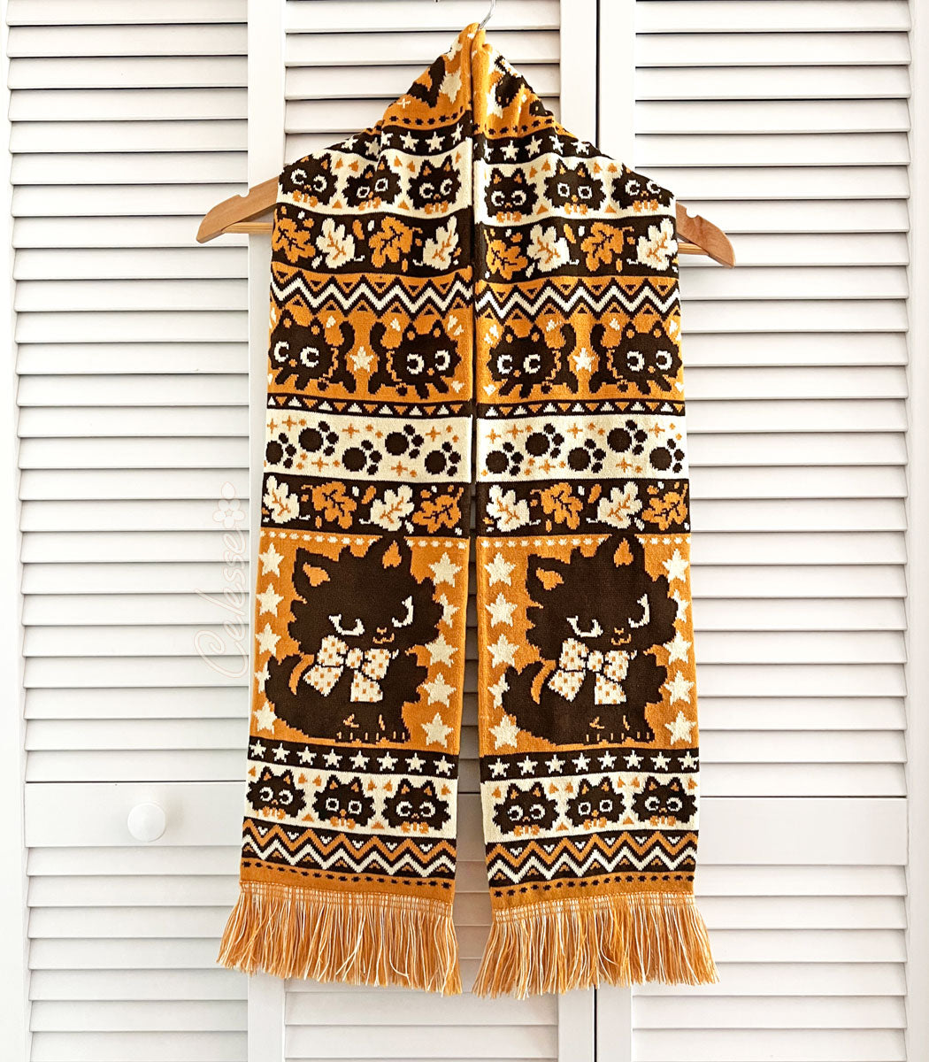 A photo of a Halloween-themed knit scarf on a wood hanger, hung on a set of white slat-walled doors. The scarf is orange, light yellow and dark brown and features rows of black cats, leaves, paw prints and other spooky motifs.
