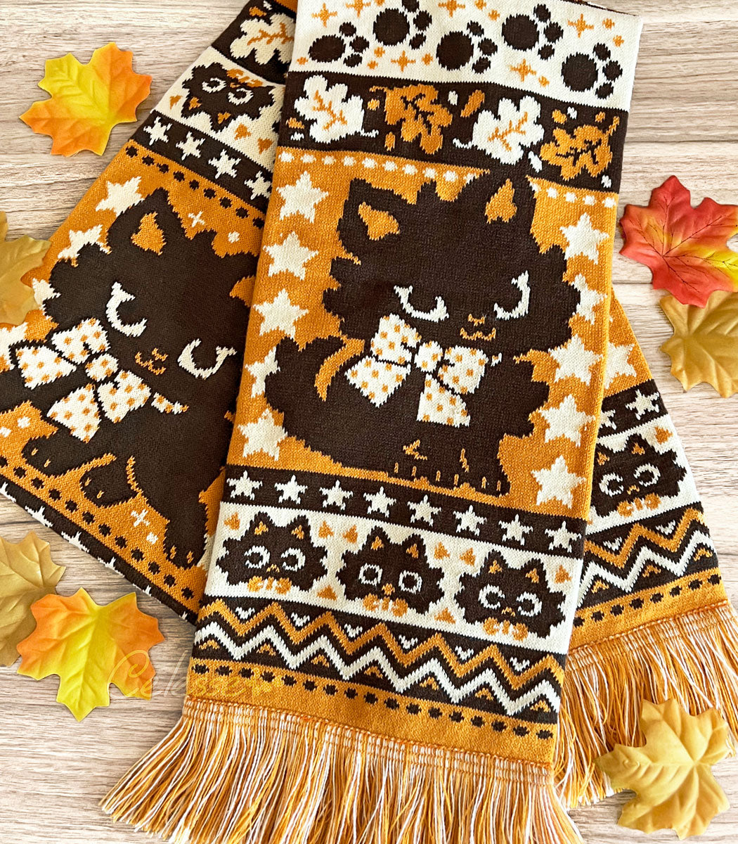 A Halloween-themed knit scarf featuring playful black cat designs on an orange background with orange and yellow fringes, displayed on a wooden surface with autumn leaves around it.