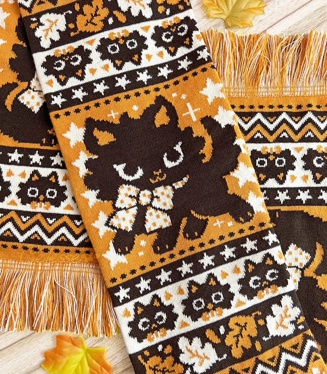 A Halloween-themed knit scarf featuring playful black cat designs on an orange background with orange and yellow fringes, displayed on a wooden surface with autumn leaves around it.