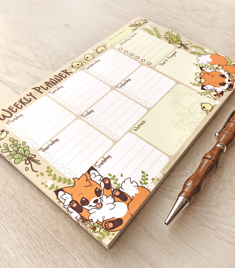 A photo of a weekly planner notepad on a wood table, the design featuring cute little foxes playing with yellow chicks, with task sections for each day of the week as well as extra notes and goals.