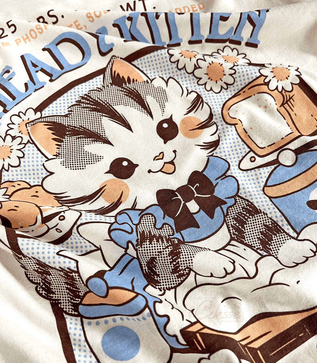 A close up photo of the print of a shirt featuring a kitten in a frilly apron kneading dough.