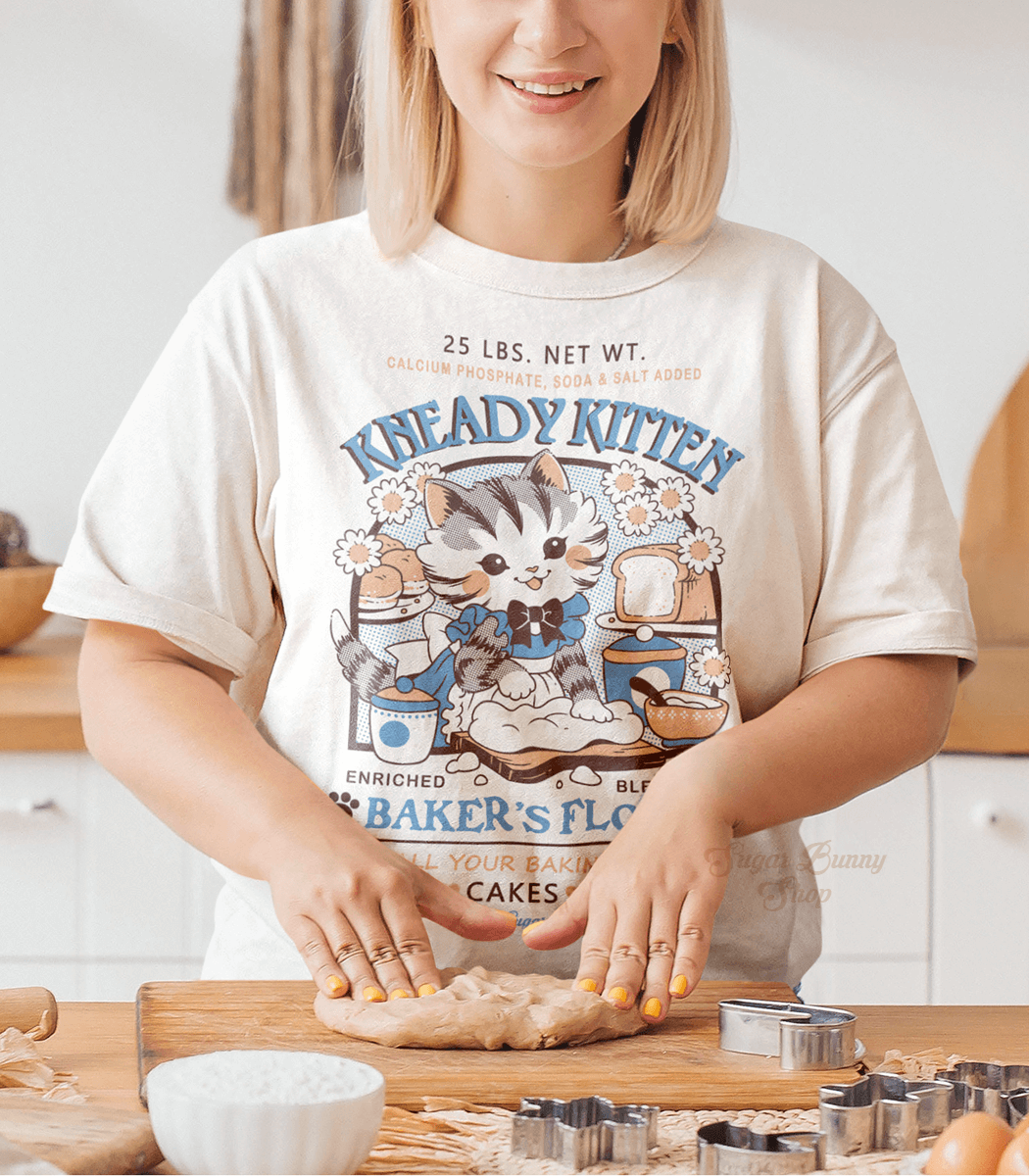 A photo of a woman smiling in a bright kitchen while kneading dough and wearing an off-white shirt with a faux vintage-style logo print for Kneady Kitten Baker&