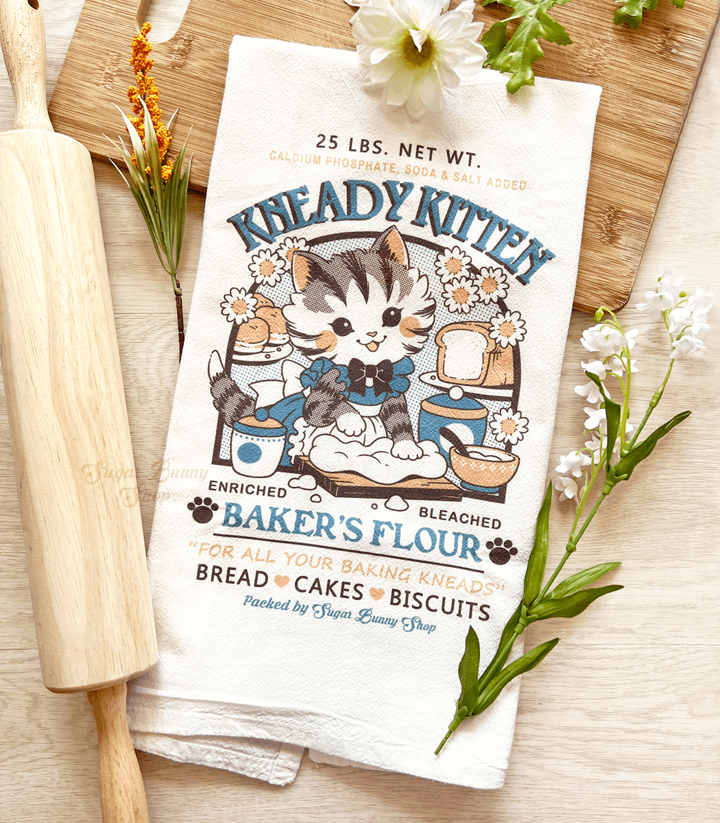 A photo of a white flour sack tea towel laid on a wood surface with a cutting board and rolling pin. The print features a vintage-style logo for Kneady Kitten Baker&