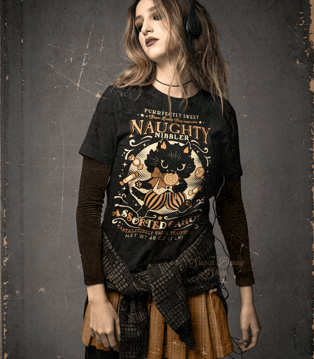A photo of a goth girl wearing a black shirt featuring a vintage-style logo in orange and yellow for Naughty Nibbler Assorted Candy. A black kitten eating a lollipop prances among various candies between the text.
