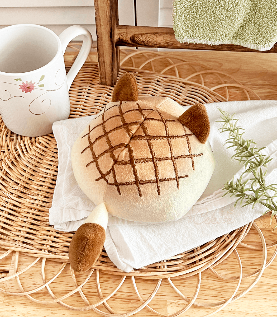 A photo of a round plush bread cat in the shape of melon bread laid out on a white towel on a kitchen counter.