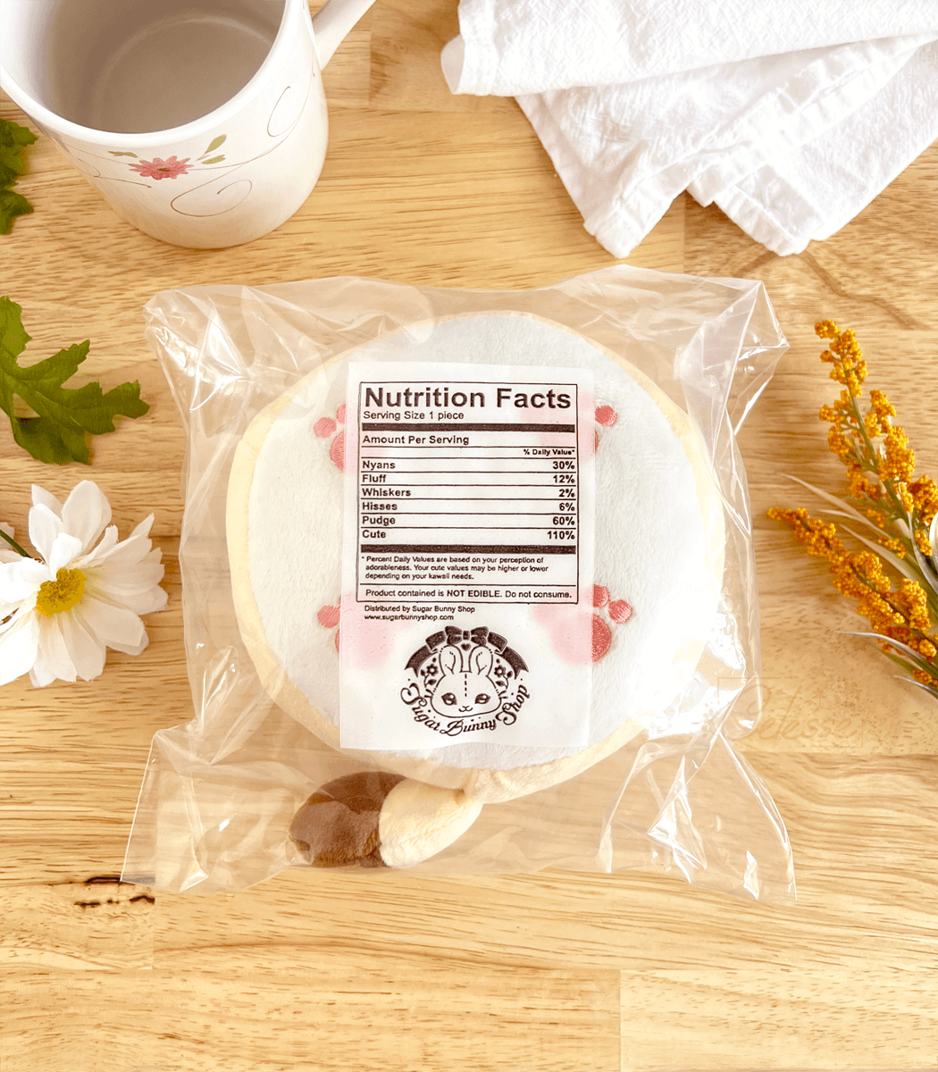 A round plush bread cat in the shape of melon bread inside plastic packaging on a kitchen counter, the underside showing a faux list of nutrition facts.