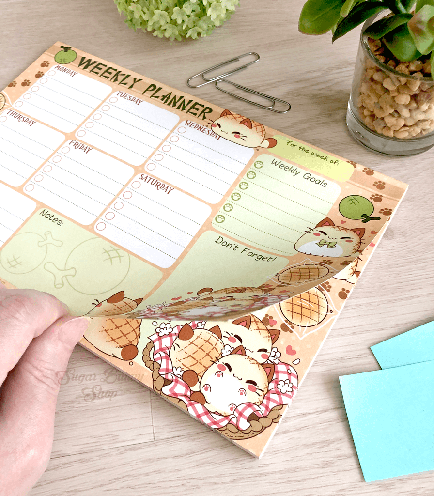 A photo of a hand lifting the page of a weekly planner notepad on a table with plants, the design featuring Nyanpan the bread cat in various poses with task sections for each day of the week, as well as extra notes and goals.