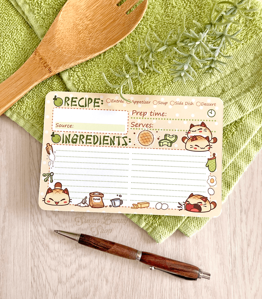 A set of recipe cards with an illustration of little bread cats and kitchen elements, placed on a green kitchen towel alongside a wooden spoon and a pen.