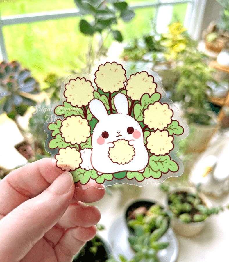 A hand holding a clear vinyl sticker in front of a table full of plants. The sticker design features a fat white bunny holding a yellow dandelion puff, with more dandelion plants spread out around them.
