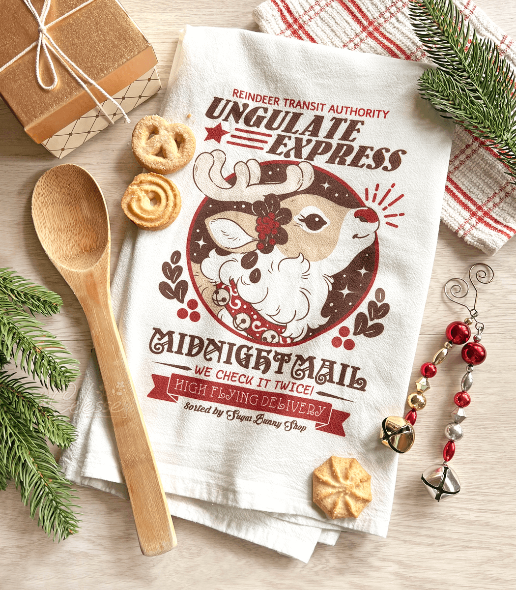 A white cotton tea towel with a vintage-style Christmas advertisement print for Ungulate Express Midnight Mail with a reindeer, displayed on a wooden surface with holiday decorations, a wooden spoon, and cookies.