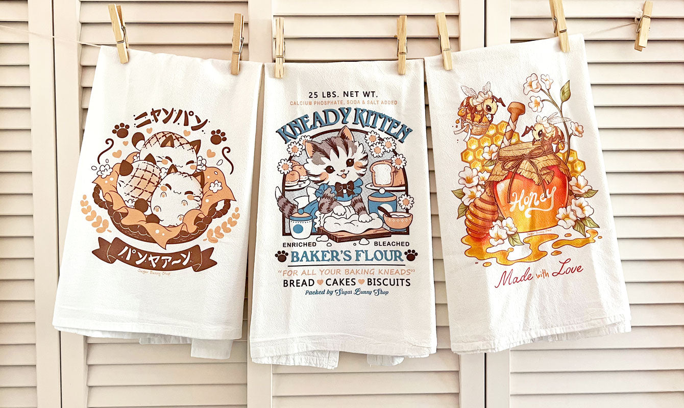 Three cotton flour sack tea towels featuring soft, vintage-style art strung up on a clothesline in front of wood slat wall door.