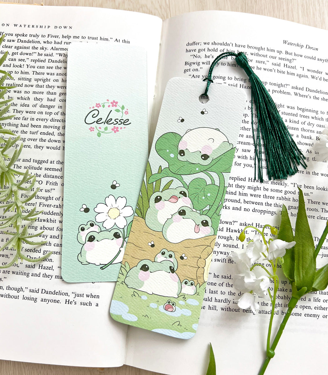 A photo of a bookmark placed atop an open book, the artwork featuring a group of fat, blob-shaped frogs gathered together on a log.