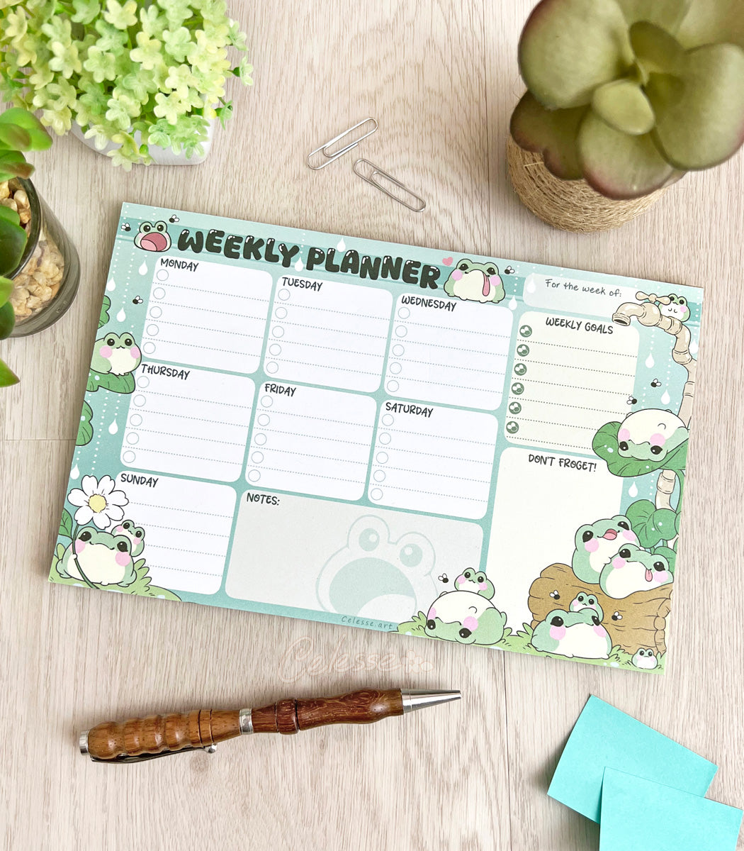 A weekly planner notepad on a wood table, the print featuring a collection of fat, blob-like frogs decorating a checklist style planner page.