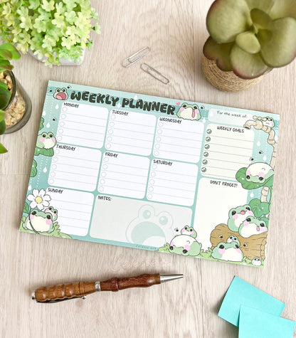 A weekly planner notepad on a wood table, the print featuring a collection of fat, blob-like frogs decorating a checklist style planner page.