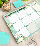 A weekly planner notepad on a wood table, the print featuring a collection of fat, blob-like frogs decorating a checklist style planner page.
