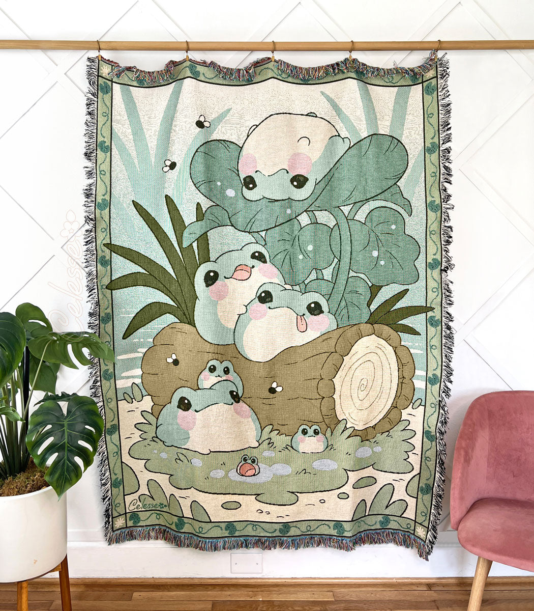 A photo of a woven tapestry throw featuring art of a group of blob-like frogs perched on logs and leaves, a border of lily pads framing the image.