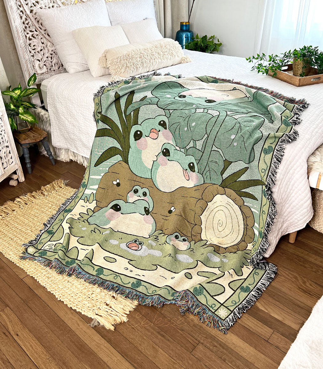 A photo of a woven blanket featuring an image of blob frogs in a pond setting. The blanket is draped off the side of a bed in a soft white bedroom with dark wood flooring.