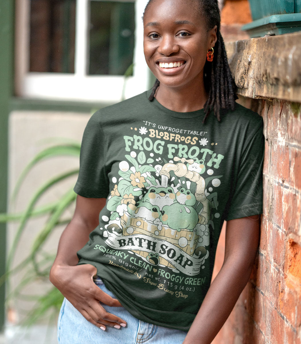 A photo of a Black woman leaning against a brick wall and wearing a dark green shirt with a print featuring a faux logo for Blobfrogs Frog Froth Bath Soap with fat frogs playing in a wooden water bucket.