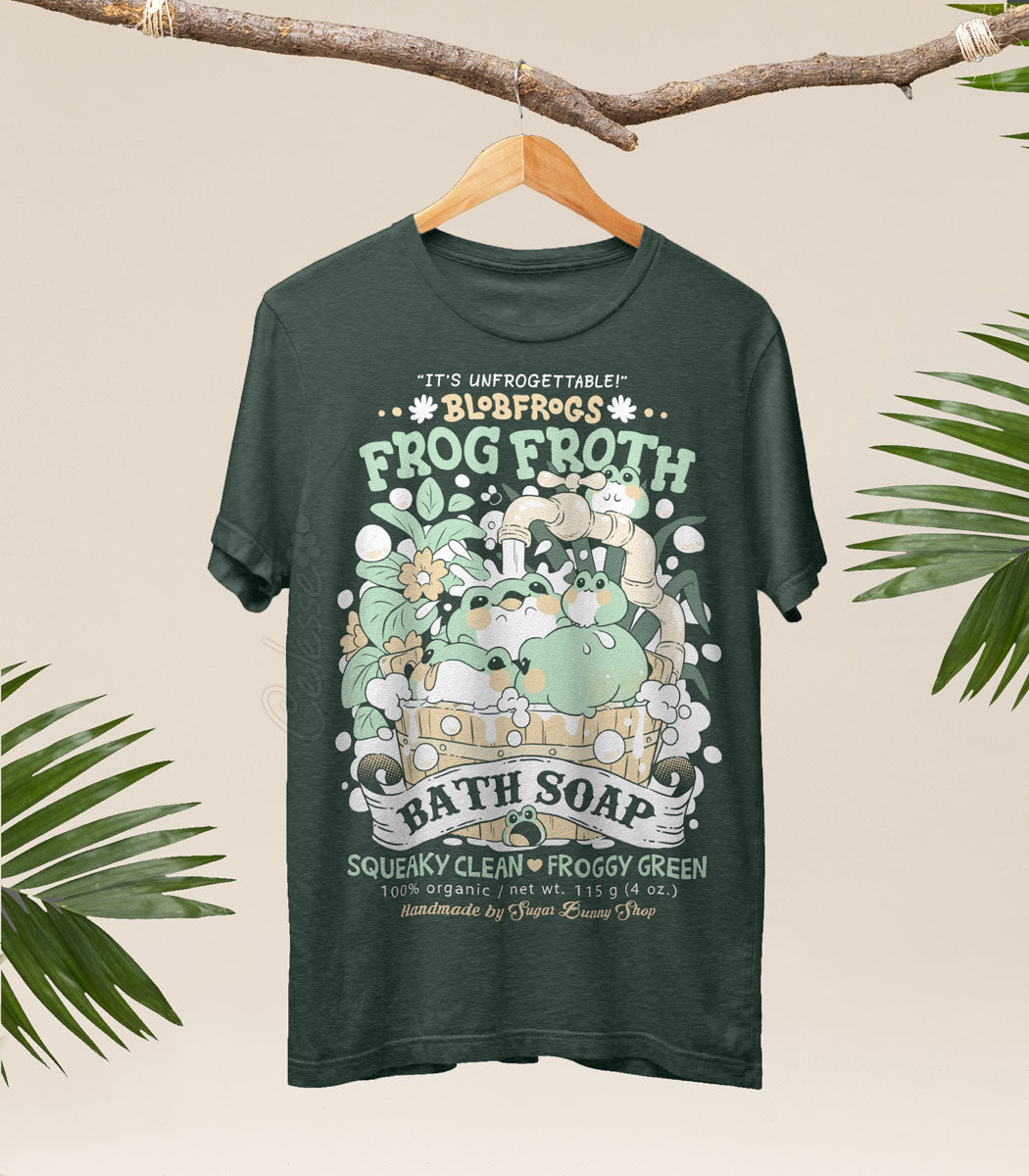 A photo of a heather forest green shirt on a wood hanger, hung from a branch with palm fronds on either side, the printed design features a vintage-style logo for Blobfrogs Frog Froth Bath Soap with a wood tub of fat squishy frogs taking a bubble bath.