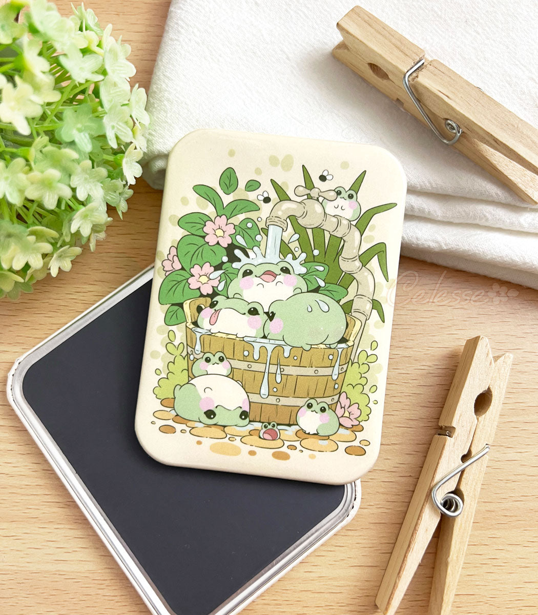 A photo of a fridge magnet laid on a table, the artwork featuring a group of blob-shaped frogs splashing around under a faucet in a wooden bucket, plants all around.
