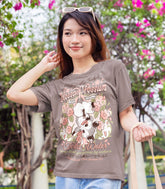A woman wearing a warm gray graphic shirt of a faux logo for Blossom Opossum Toilet Water, standing outdoors in front of flowering trees. The printed logo features a baby opossum hanging upside down among flowers.