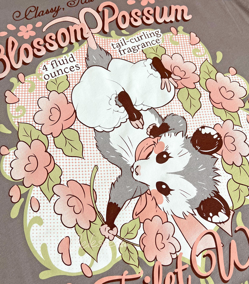 A close-up photo of the print of a warm gray shirt with a vintage-style logo featuring a hanging baby opossum surrounded by flowers.