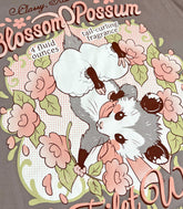 A close-up photo of the print of a warm gray shirt with a vintage-style logo featuring a hanging baby opossum surrounded by flowers.