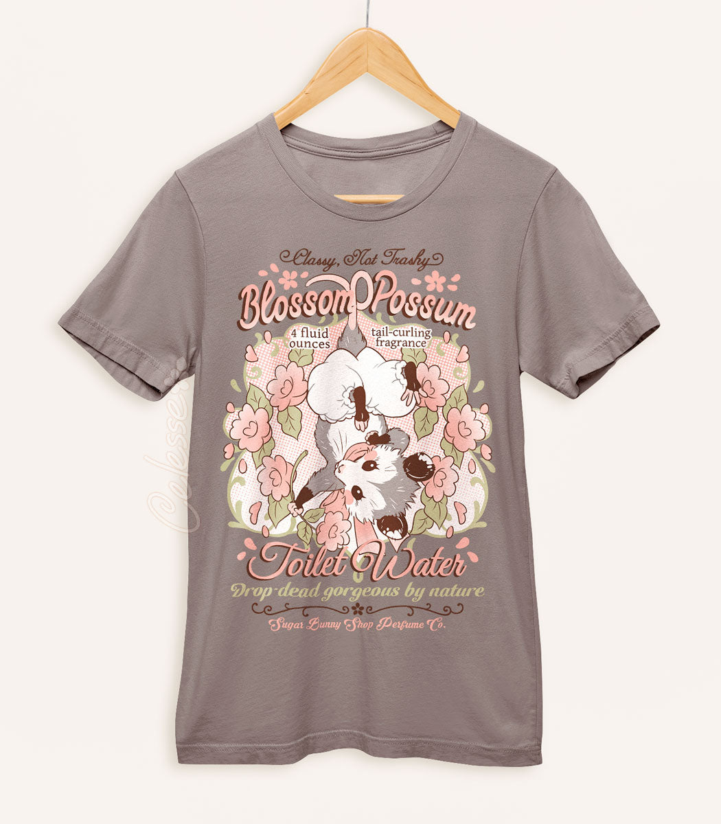 A warm gray shirt on a wood hanger with a print of a vintage-style logo for Blossom Opossum Toilet Water, the art featuring a baby opossum in bloomers hanging upside down and surrounded by blossoms. The flavor text reads "Classy, Not Trashy" and "Drop-dead gorgeous by nature."