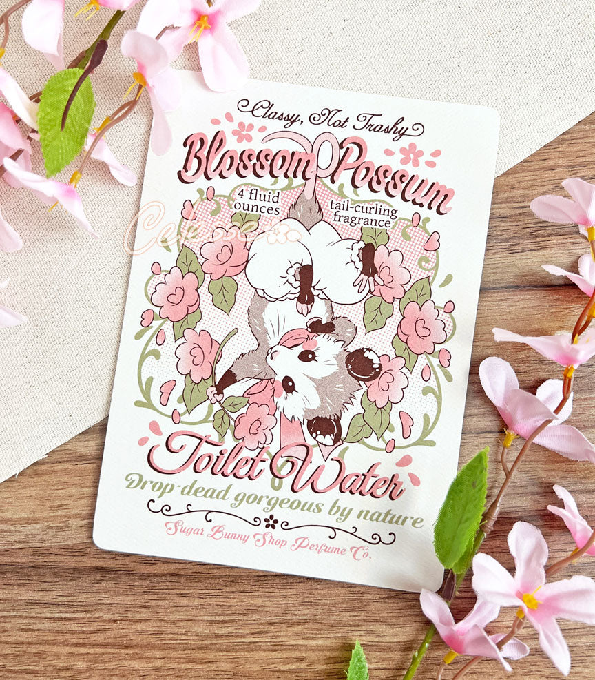 An art print of a vintage-style logo for Blossom Opossum Toilet Water, the art featuring a baby opossum in bloomers hanging upside down and surrounded by blossoms. The flavor text reads "Classy, Not Trashy" and "Drop-dead gorgeous by nature."