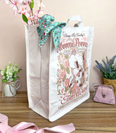 A side view of a white canvas tote bag with a whimsical vintage-style logo for Blossom Opossum Toilet Water, styled on a wooden surface with decorative flowers, a pink purse, and a floral scarf.