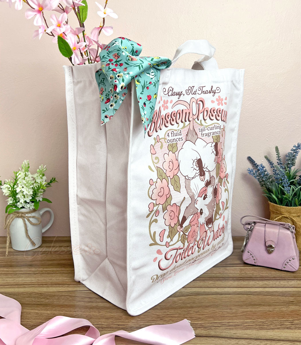 A side view of a white canvas tote bag with a whimsical vintage-style logo for Blossom Opossum Toilet Water, styled on a wooden surface with decorative flowers, a pink purse, and a floral scarf.