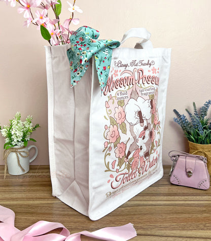 A side view of a white canvas tote bag with a whimsical vintage-style logo for Blossom Opossum Toilet Water, styled on a wooden surface with decorative flowers, a pink purse, and a floral scarf.