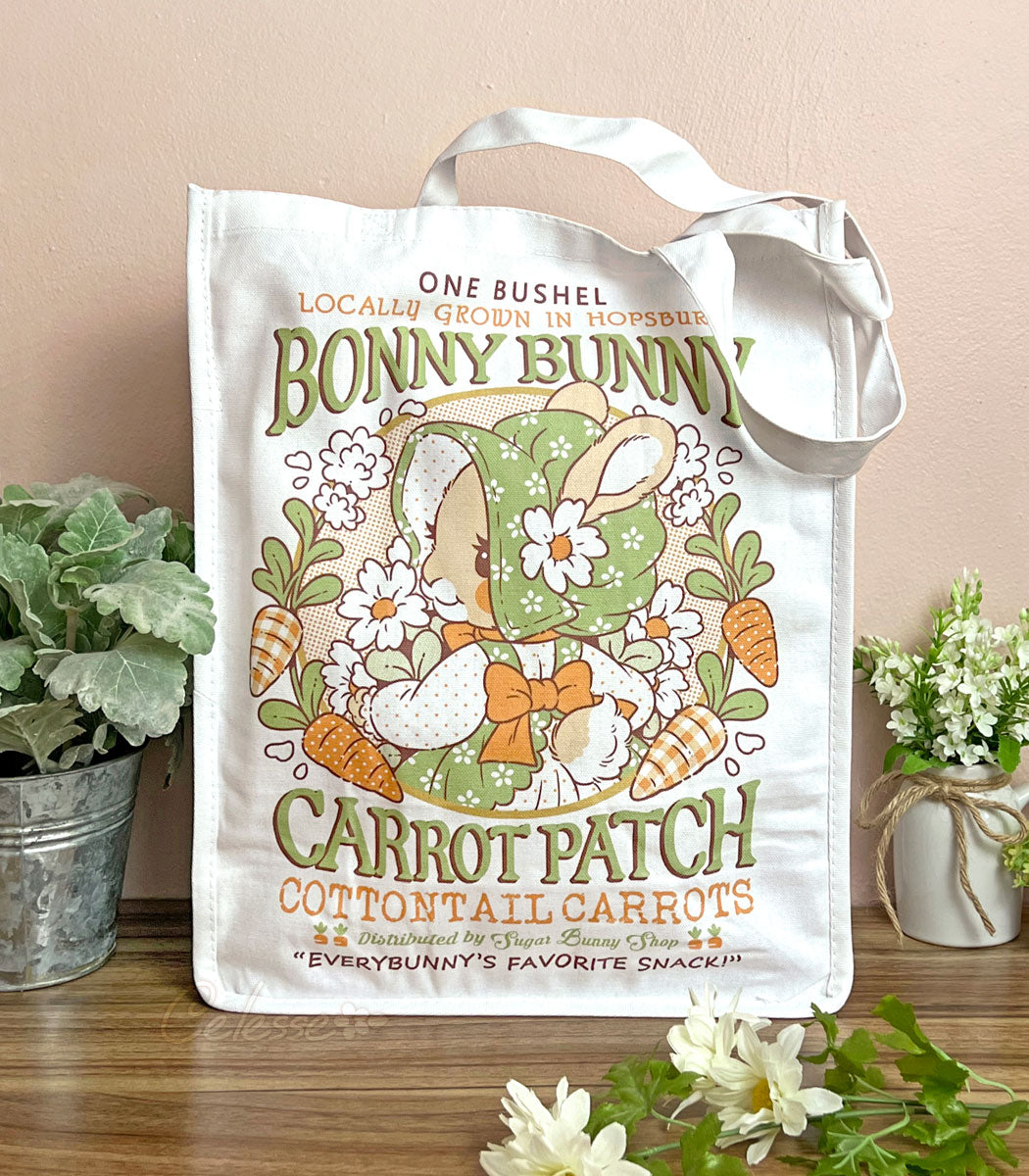 A photo of a white canvas tote on a table against a warm pink wall, plants and flowers on either side of it. The front of the bag features a print of a faux vintage-style logo for Bonny Bunny Carrot Patch, with a bunny in a bonnet holding a bundle of flowers and carrots around her.