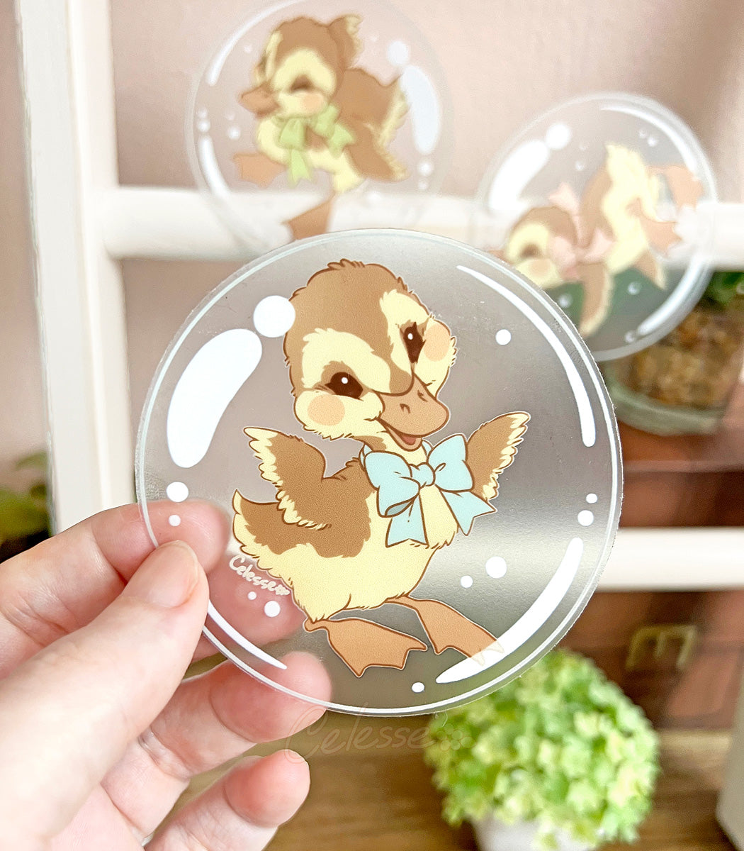 A photo of a hand holding a clear vinyl sticker with artwork of a happy duckling floating in a bubble, with more stickers and some plants in the background.