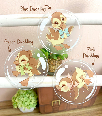 A photo of three clear vinyl stickers featuring artwork of ducklings floating inside bubbles, displayed on a white wood stand with a neutral background.
