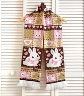 A knit scarf with a patchwork bunny pattern in Neapolitan colors hanging on a white slat wall background.