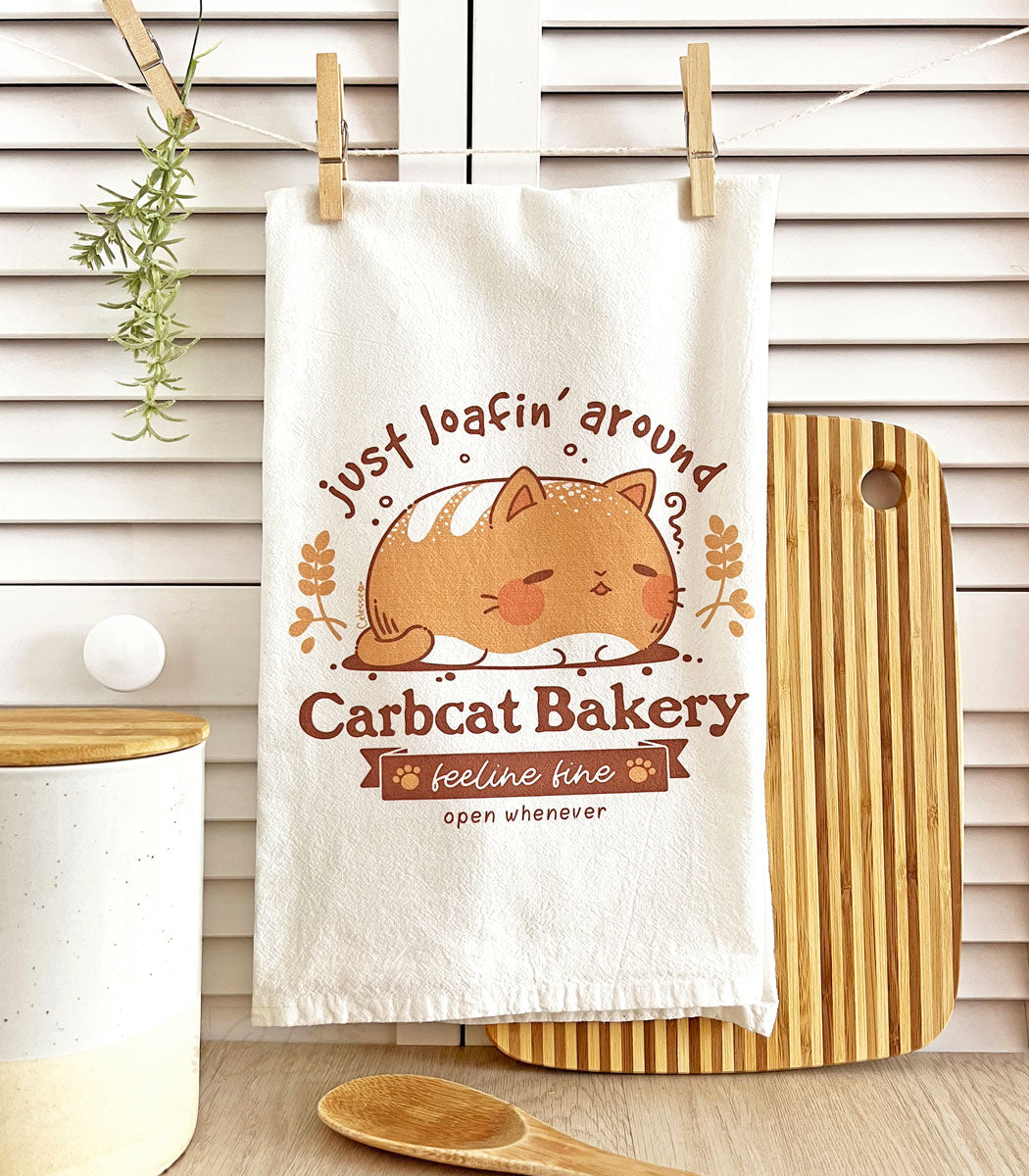 A photo of a white tea towel featuring a bread loaf cat logo called Carbcat Bakery, hung on a line with clothespins in front of a white shutter. A lined cutting board and a canister are on either side.