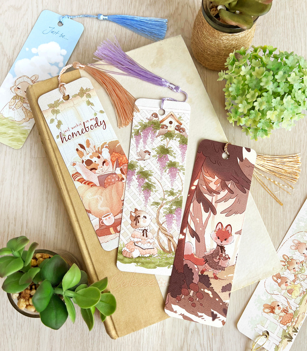 Set of colorful bookmarks with tassels laid out on top of a book atop a wooden surface with plants around it.