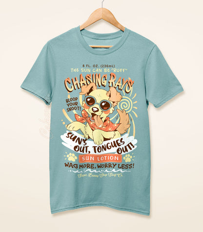 A dusty blue shirt on a wood hanger with a print of a vintage-style logo for Chasing Rays Sun Lotion, the art featuring an energetic, floppy-eared dog wearing a bandana and sunglasses. The flavor text reads, "Sun&