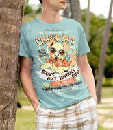 A man wearing a light blue graphic t-shirt with a cartoon dog and text about sun lotion, standing outdoors on a beach next to a palm tree trunk.