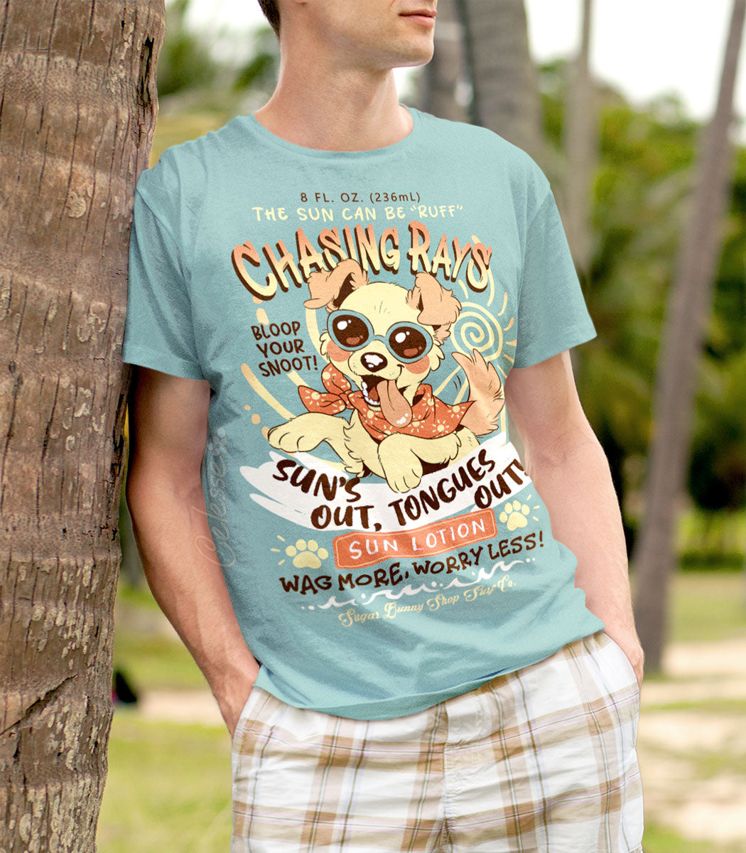 A man wearing a light blue graphic t-shirt with a cartoon dog and text about sun lotion, standing outdoors on a beach next to a palm tree trunk.