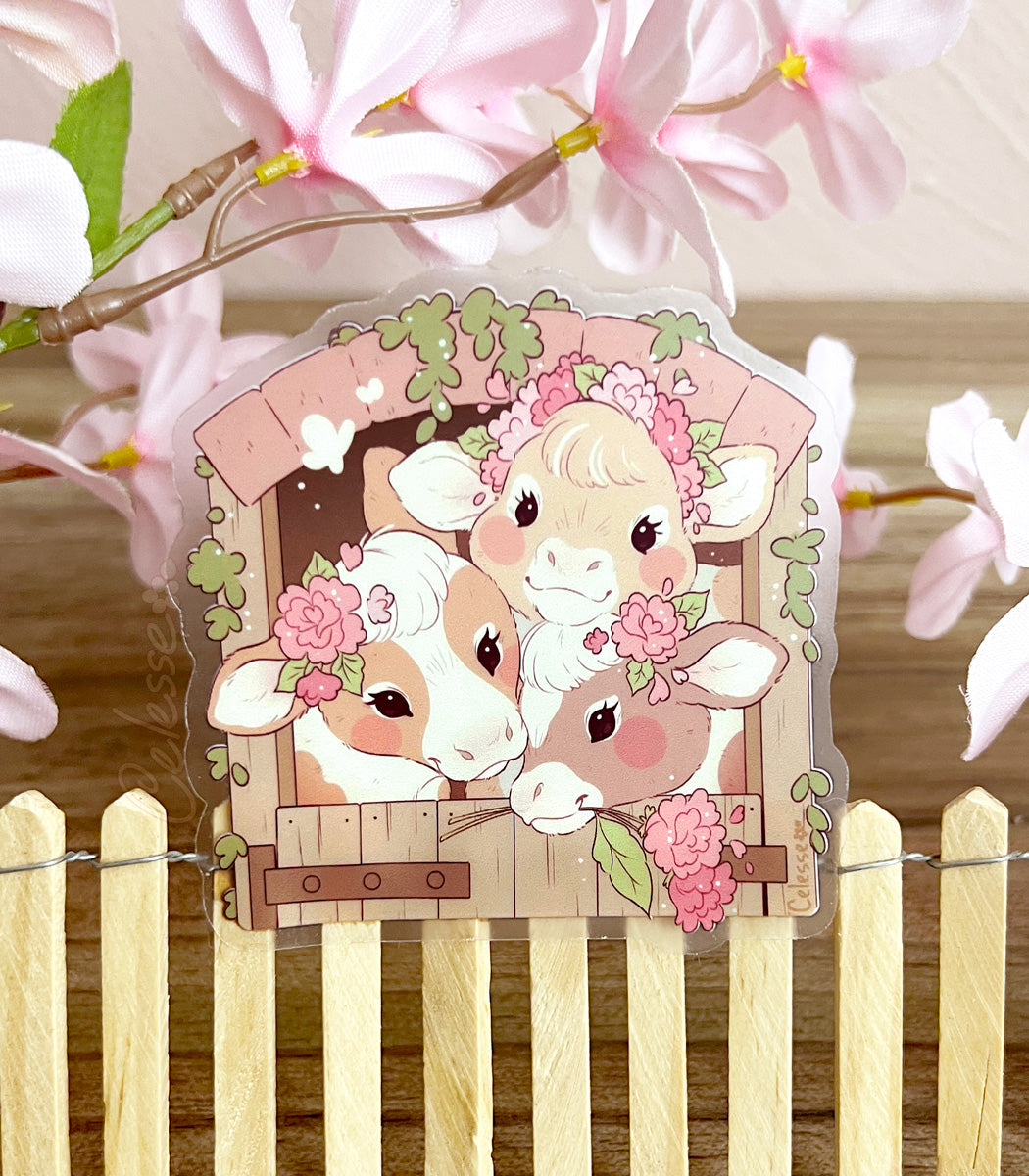 A photo of a clear vinyl sticker featuring artwork of three country cows in a barn, stuck atop a mini wooden fence with pink flowers in the background.