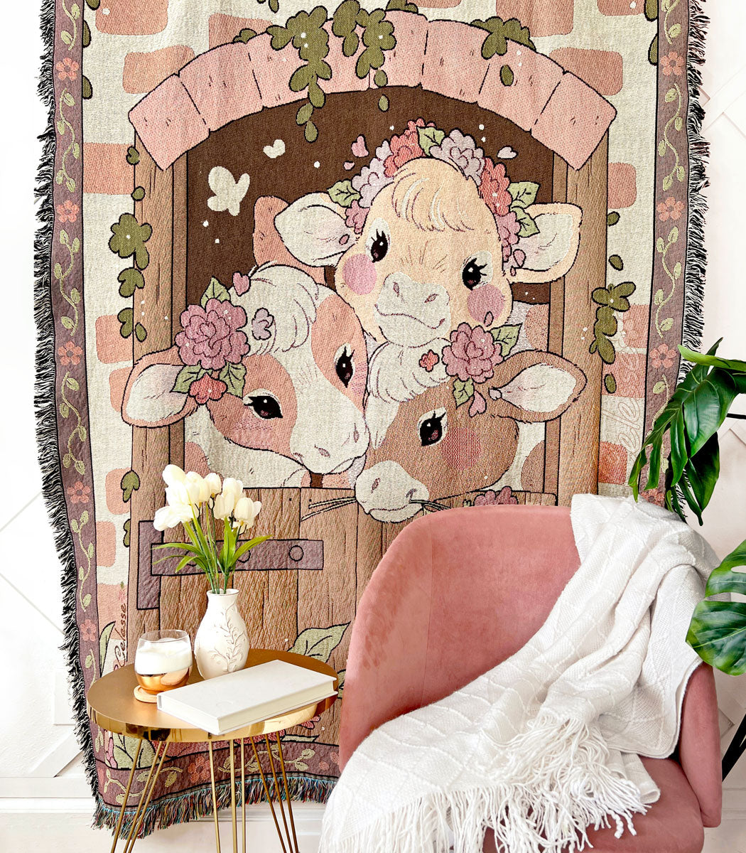 A photo of a woven tapestry blanket hung on a wall with a chair and small table with items on it positioned in front. The blanket features 3 country cows looking through a barn door set in a brick wall.