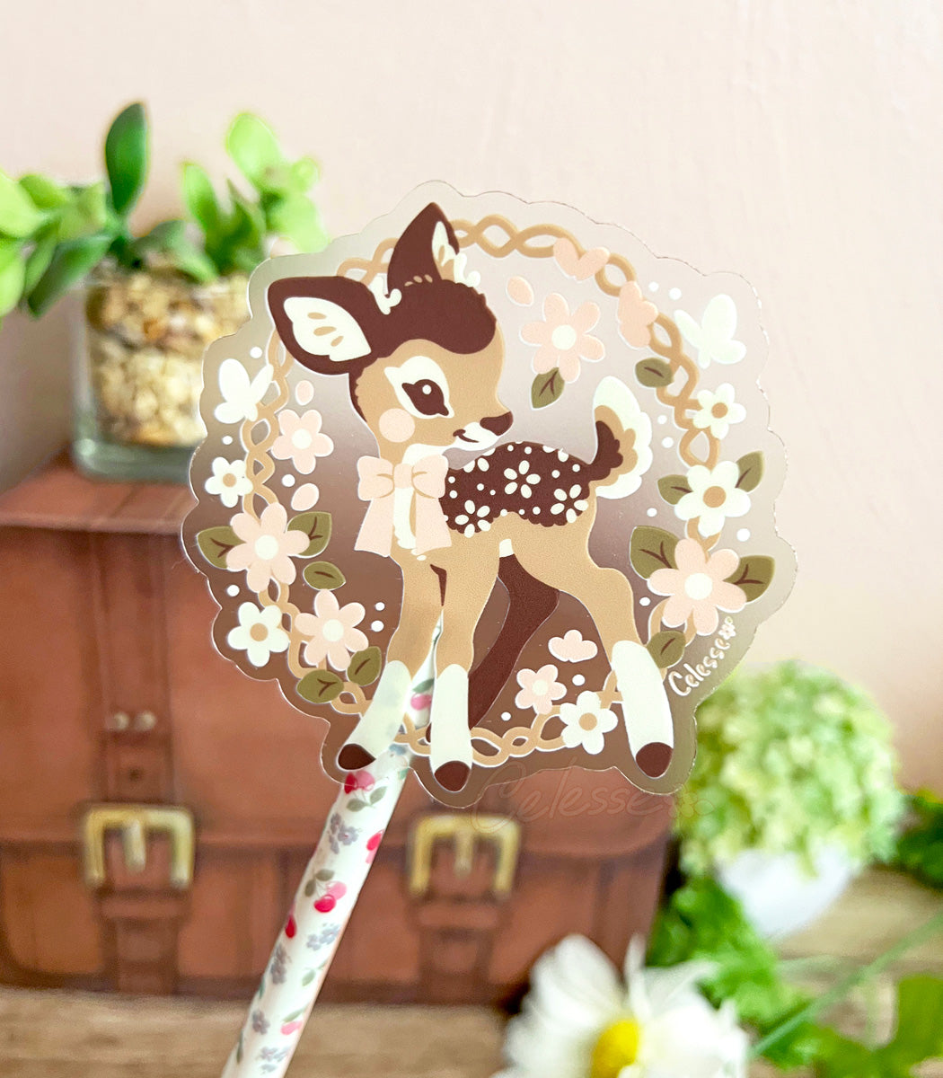 A photo of a decorative clear vinyl sticker of a baby deer with flower spots standing inside a floral wreath, set against a blurred indoor background.