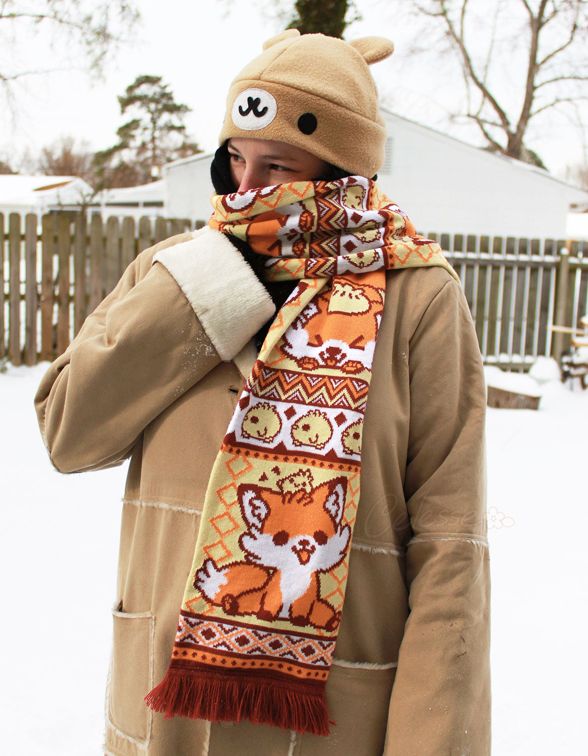 A photo of a person in a snowy landscape with a wood fence in back, wearing a light brown coat and a fleece bear hat, a colorful orange, yellow and red fox-themed knit scarf wrapped around their neck.