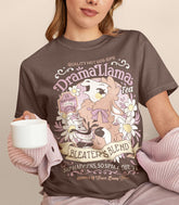 A woman holding a cup and wearing a brown shirt with a print for a faux vintage-style logo for Drama Llama Tea, the art featuring a gossipy llama spilling the tea.